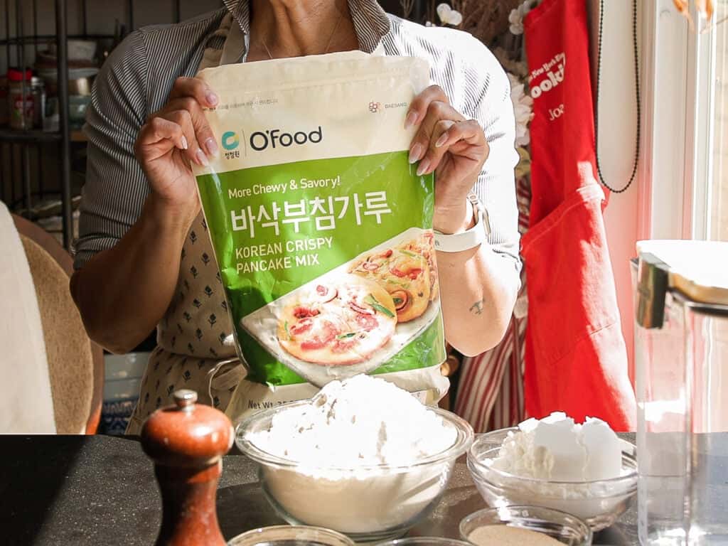 A person in an apron holds a bag of OFood Korean Crispy Pancake Mix over a kitchen counter with bowls of flour, a pepper grinder, and kitchen tools in sunlight.